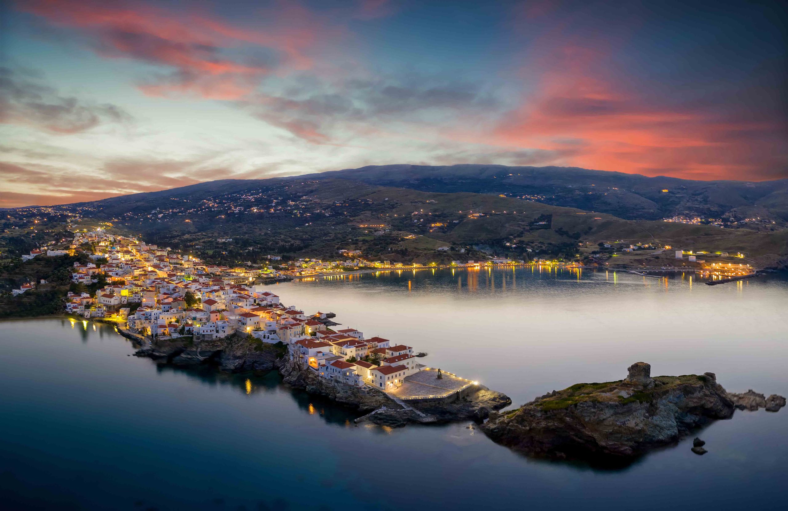 Aerial sunset view of the beautiful illuminated town of Andros island, Cyclades, Greece, set on rocks reaching out into the sea